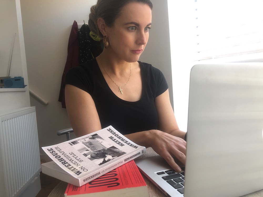 News Associates Manchester part-time trainee Gemma Corby wearing a black t-shirt with her dark blonde hair tied back and hoop earrings sitting at a desk on a laptop next to a pile of two books: Waterhouse on Newspaper Style by Keith Waterhouse and The Universal Journalist by David Randall