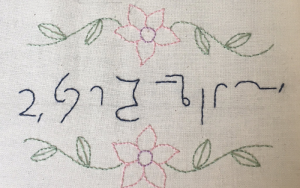 The sentence 'Good evening ladies and gentlemen and welcome to this parish council meeting' in Teeline shorthand sewn into a white cloth in blue thread surrounded by sewn in green leaves and light pink flowers.