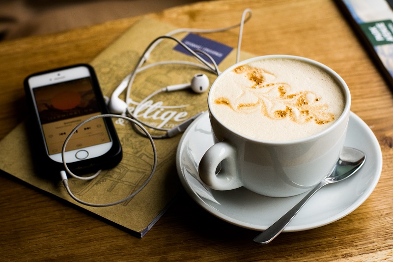 iPhone and coffee mug stock photo. White iphone is on the left, showing a podcast or song being played. White earphones are lying on top of a brown coffee shop menu, with a creamy latte in a mug on the right, sitting on a saucer with a teaspoon next to it. The objects are placed on a wooden table.