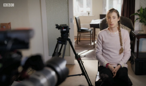 Screenshot from Panorama. Camera in foreground to the left, girl in pink jumper sitting on a chair to the right being filmed in a house.