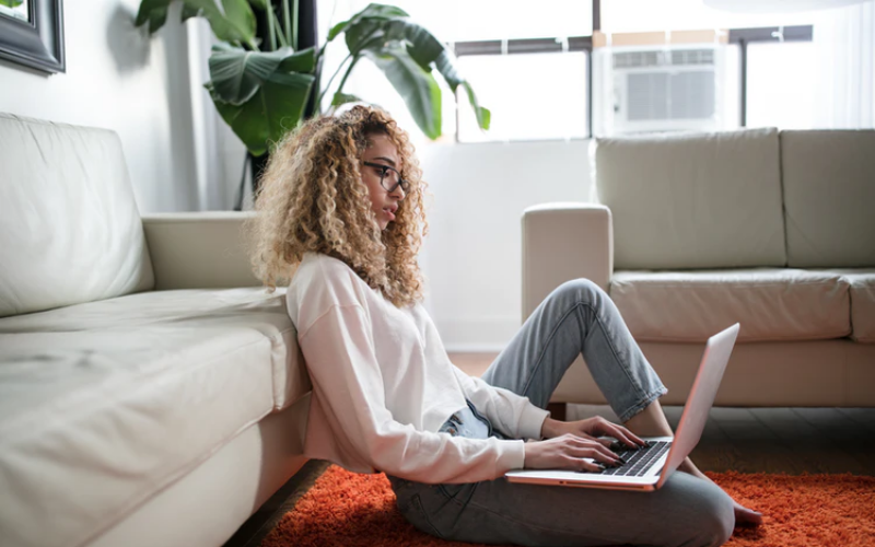 Stock image of young woman sat on the floor of a living room typing on a laptop. She's got blonde curly hair and is wearing a white shirt and blue jeans. There is a lare houseplant behind her, an orange rug and two cream sofas.