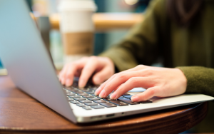 Stock photo of someone typing on a laptop, wooden desk, person is wearing a green jumper
