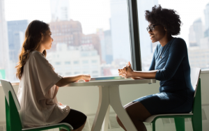 Stock image of two women chatting at an office table. Big window behind them, both women are resting their hands on the table and smiling.