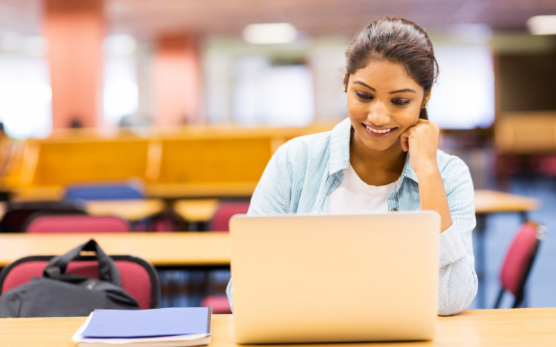 Stock photo of a young woman smiling and looking at a laptop. Sat in a classroom/library, wearing a pale blue shirt.