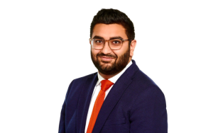 Photo of Inzamam Rashid wearing a suit with a white shirt and a red tie, smiling, wearing glasses with a white background