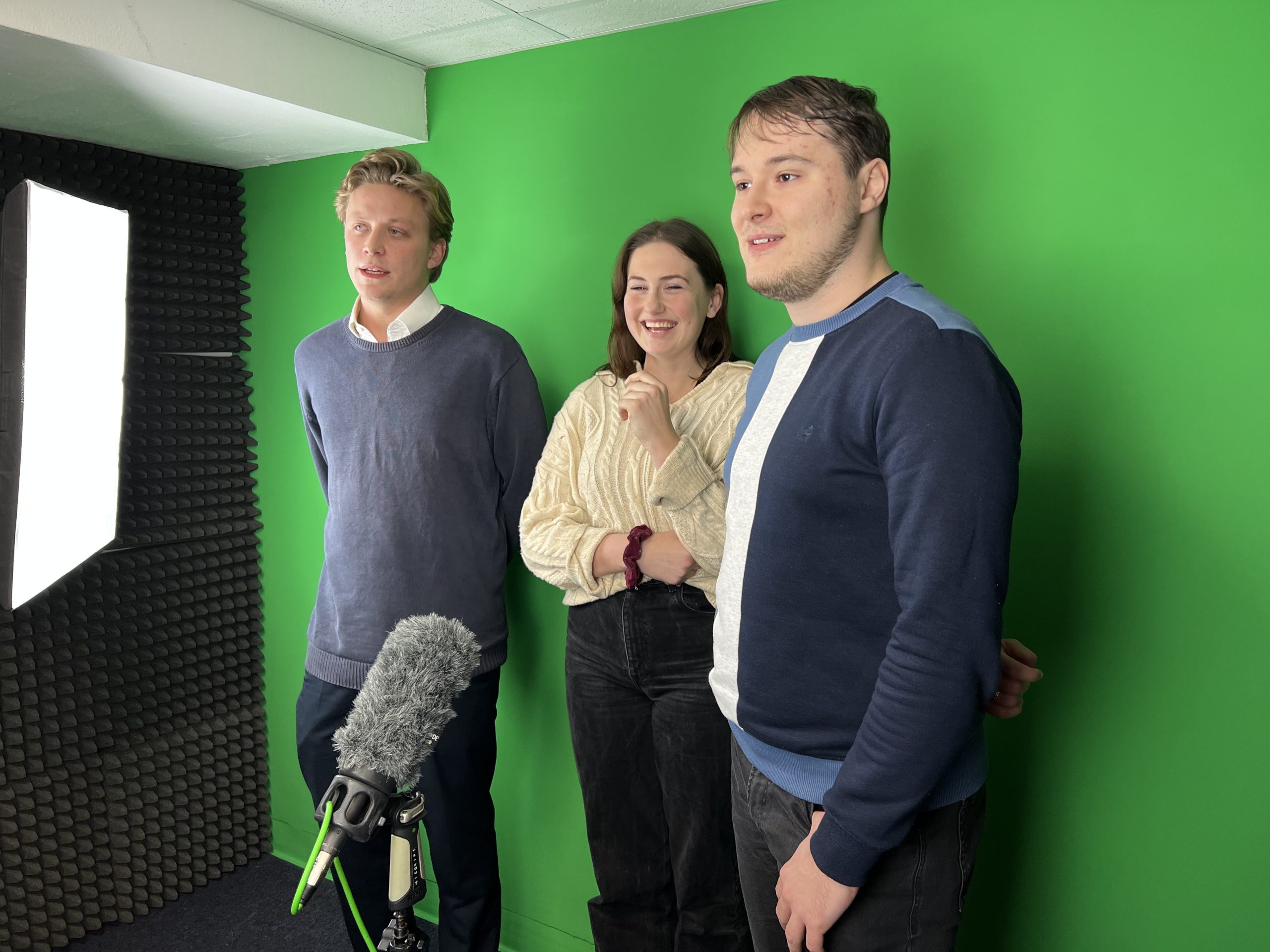 Three trainees are standing in front of a green screen and microphone in the School of Journalism studio.