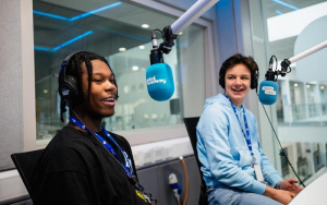 An image supplied from the Global Academy. Two students are in a radio studio at the Global Academy. The students are talking and laughing into blue Global Academy branded microphones.