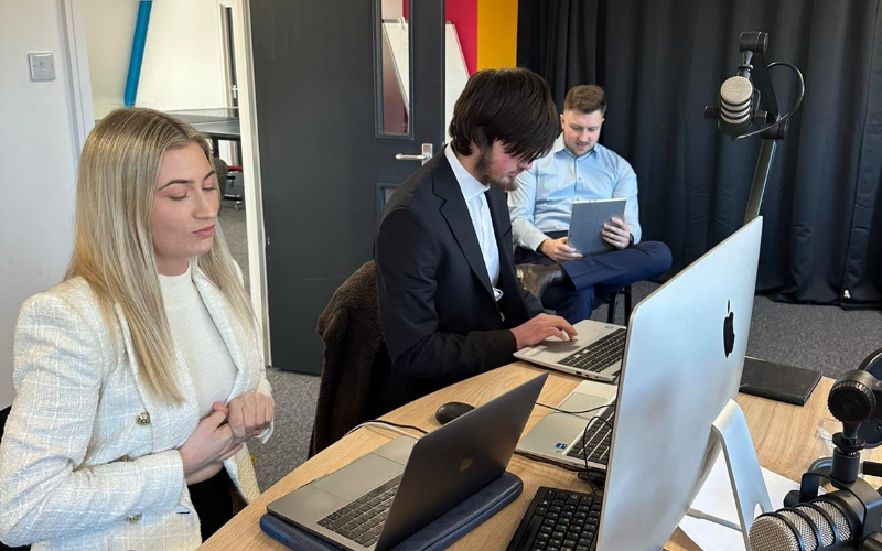 A group of three School of Journalism trainees dressed smartly prepare for a broadcast task with laptops and iPads to hand, in a broadcast studio with black curtains and microphones.