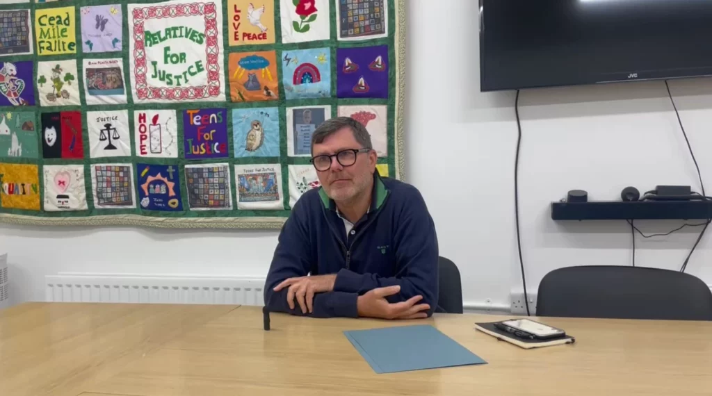 A photograph of Mark Thompson being interviewed by Emma Butterworth sat at a desk with a TV behind him for her documentary on the troubles in Northern Ireland