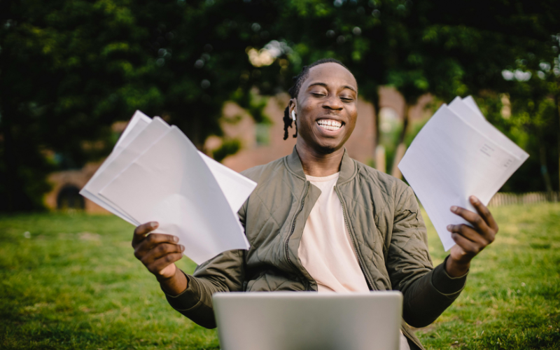 A photo of someone smiling at their laptop and sheets of paper. We are using this image to illustrate an article about A Level Results Day 2025.