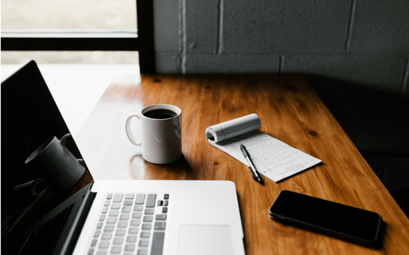A photo of a coffee, notepad and laptop on a table.